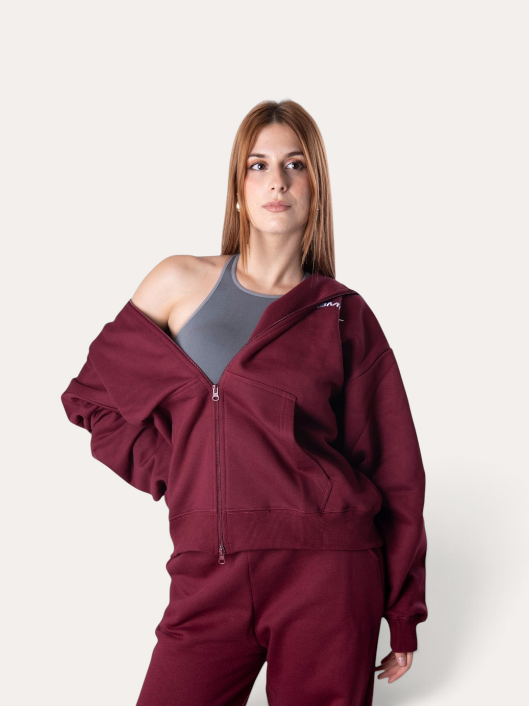Modella indossa la felpa con cappuccio Bombafit Flux color bordeaux con zip frontale sopra un top sportivo grigio