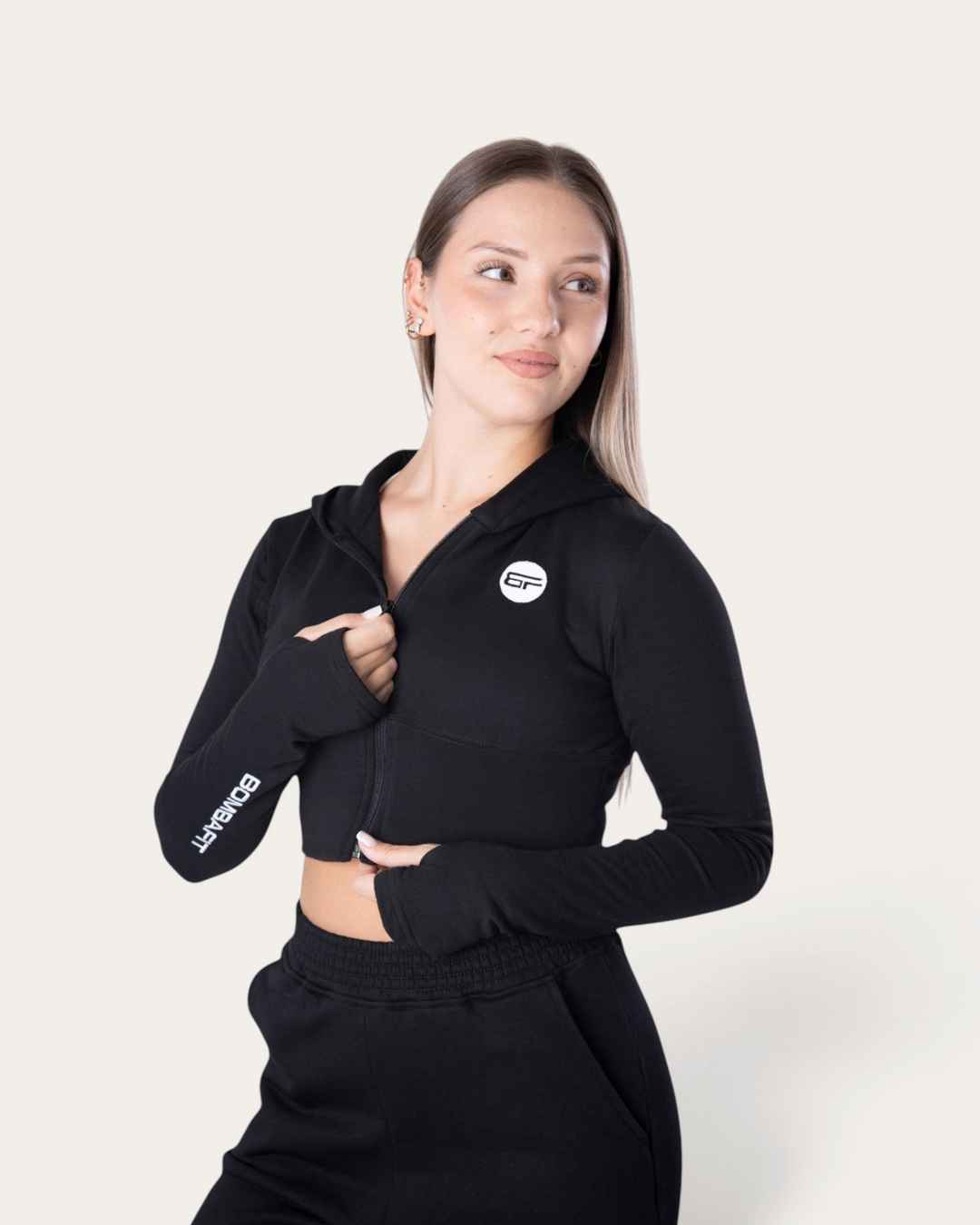 Modella indossa la felpa corta con zip Bombafit Flux Mini nera con fori per i pollici e logo sul petto