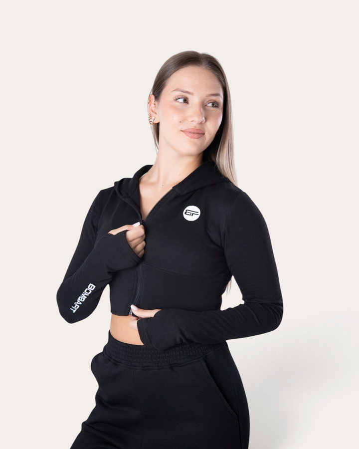 Modella indossa la felpa corta con zip Bombafit Flux Mini nera con fori per i pollici e logo sul petto