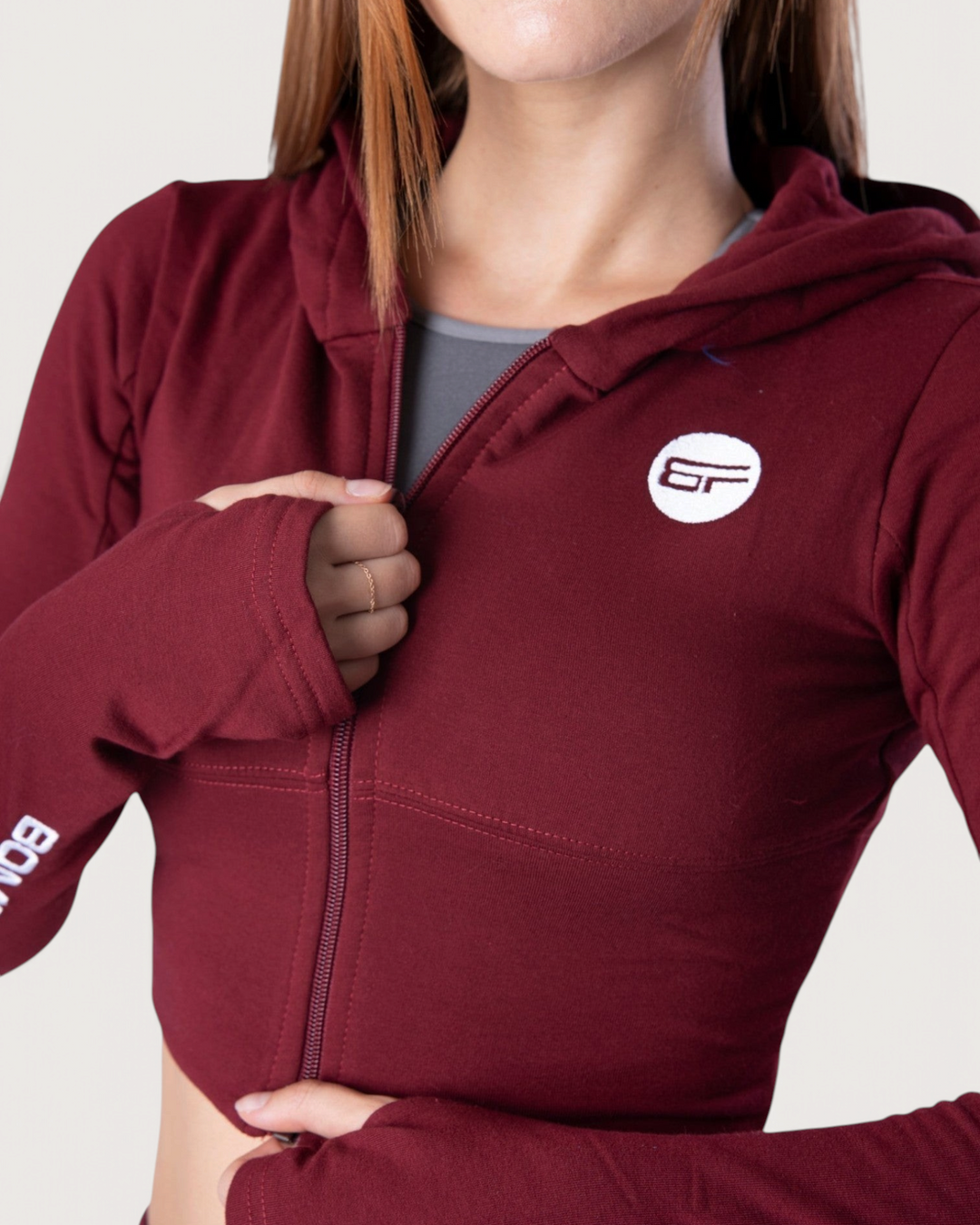 Donna che indossa la felpa con cappuccio Bombafit Flux Mini color bordeaux con zip frontale e fori per i pollici