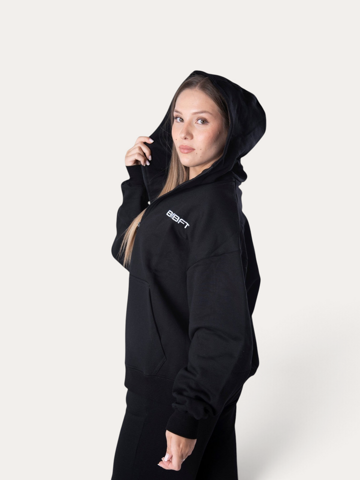 Modella indossa la felpa con cappuccio nera Flux Hoodie di Bombafit con logo bianco sul petto
