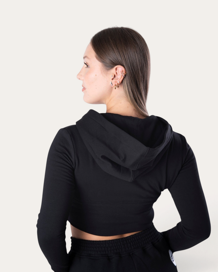 Modella indossa la felpa corta nera Bombafit Flux Mini con cappuccio, vista posteriore su sfondo neutro