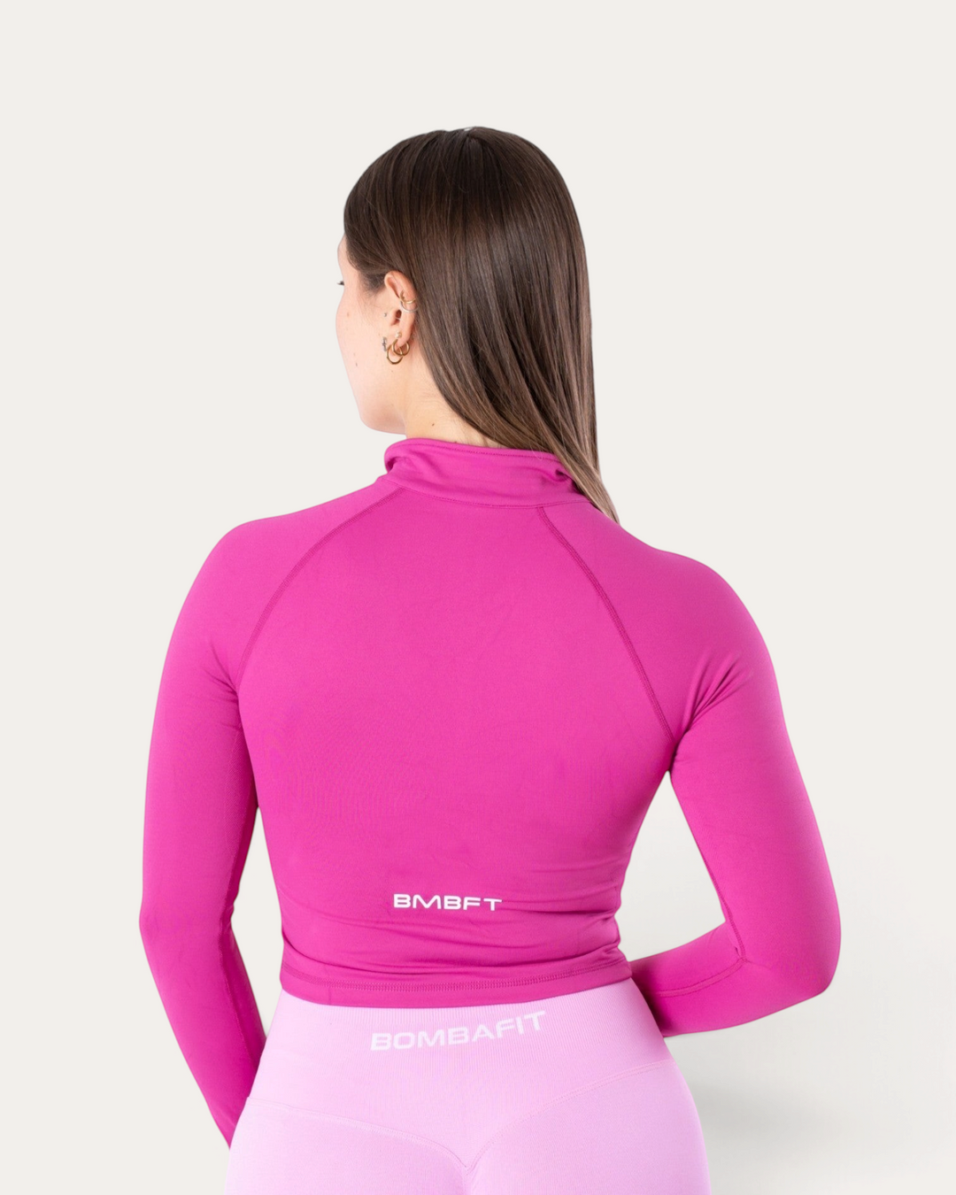 Vista posteriore di una giacca sportiva a maniche lunghe Bombafit color fucsia con zip e logo BMBFT sulla schiena