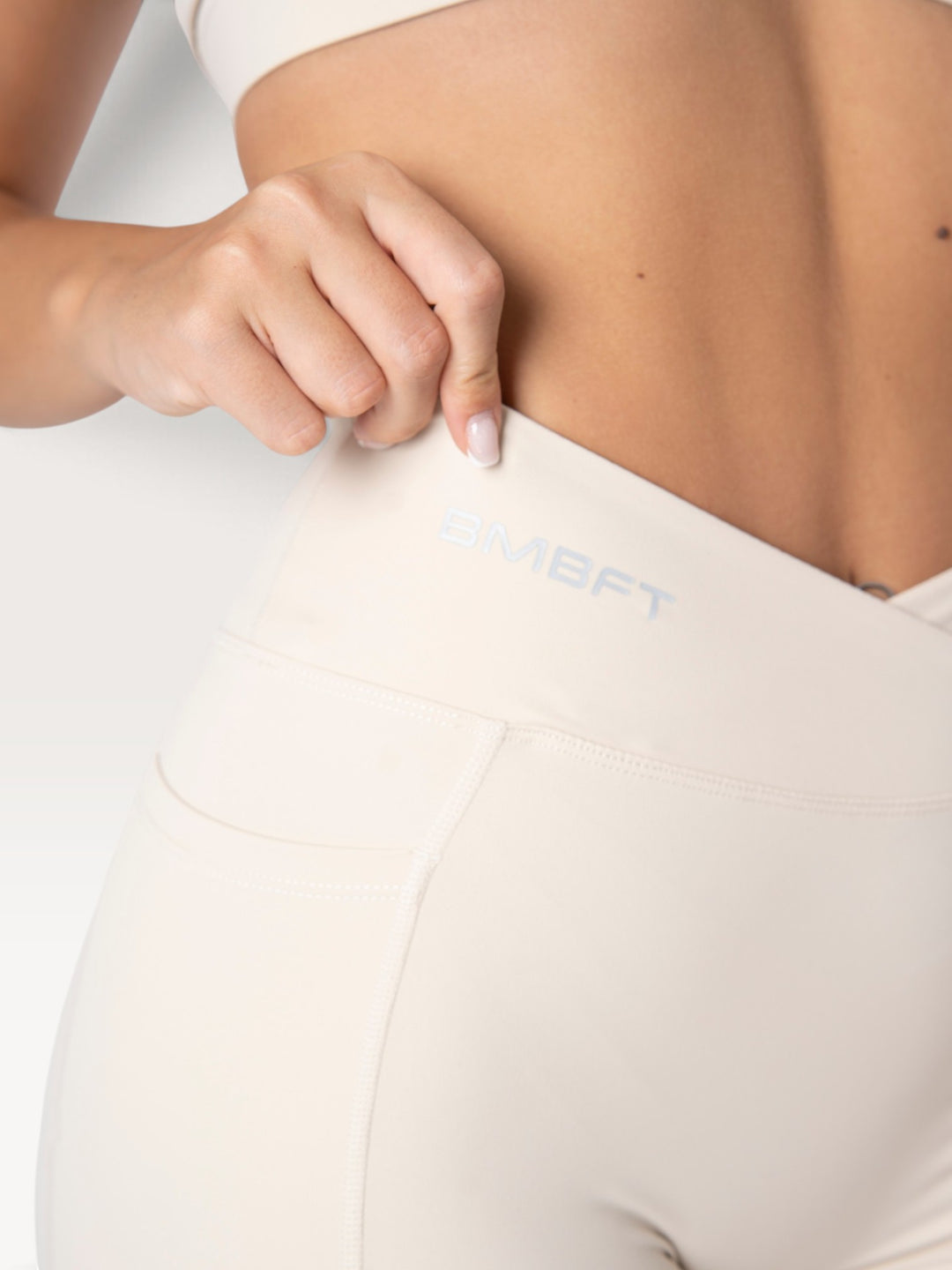 Dettaglio dei leggings Bombafit beige a vita alta con incrocio frontale, tasca laterale e logo BMBFT stampato.