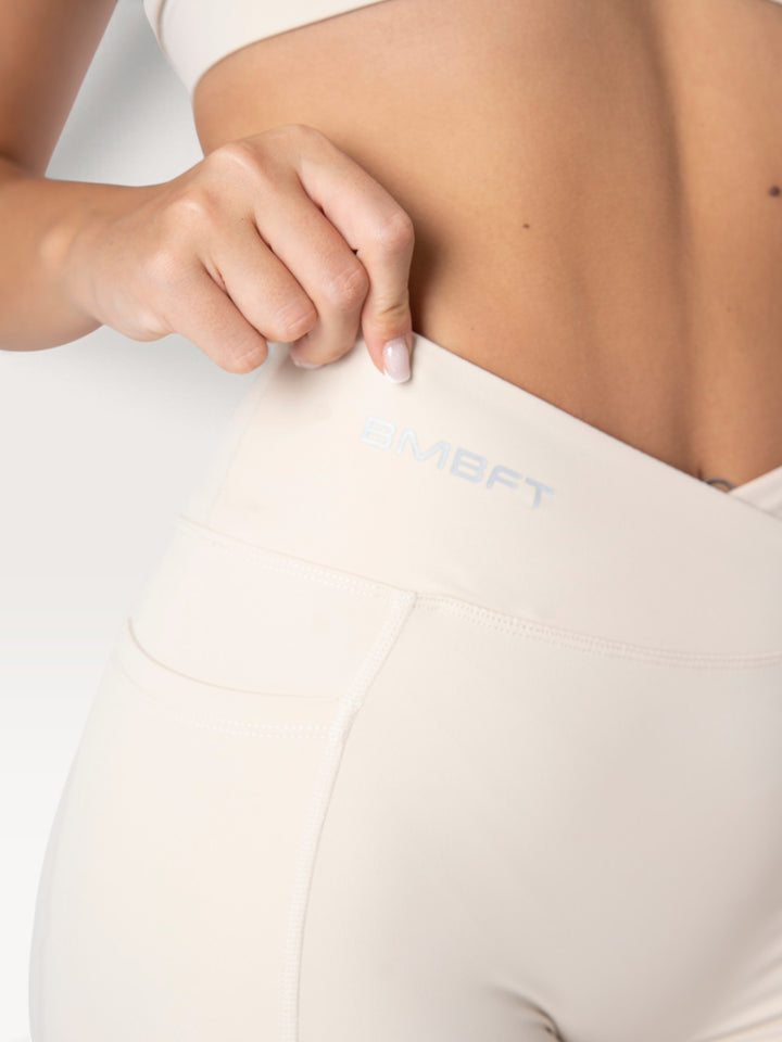 Dettaglio dei leggings Bombafit beige a vita alta con incrocio frontale, tasca laterale e logo BMBFT stampato.