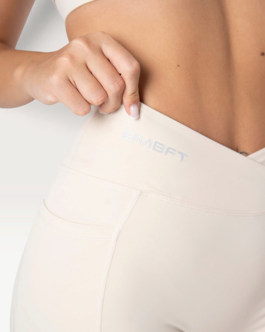 Dettaglio dei leggings Bombafit beige a vita alta con incrocio frontale, tasca laterale e logo BMBFT stampato.