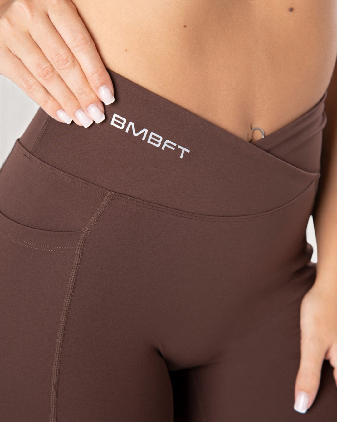 Dettaglio vita alta incrociata dei leggings Bombafit marroni con logo BMBFT e tasca laterale per fitness