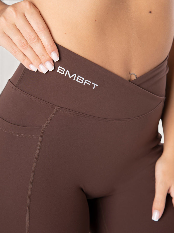 Dettaglio vita alta incrociata dei leggings Bombafit marroni con logo BMBFT e tasca laterale per fitness