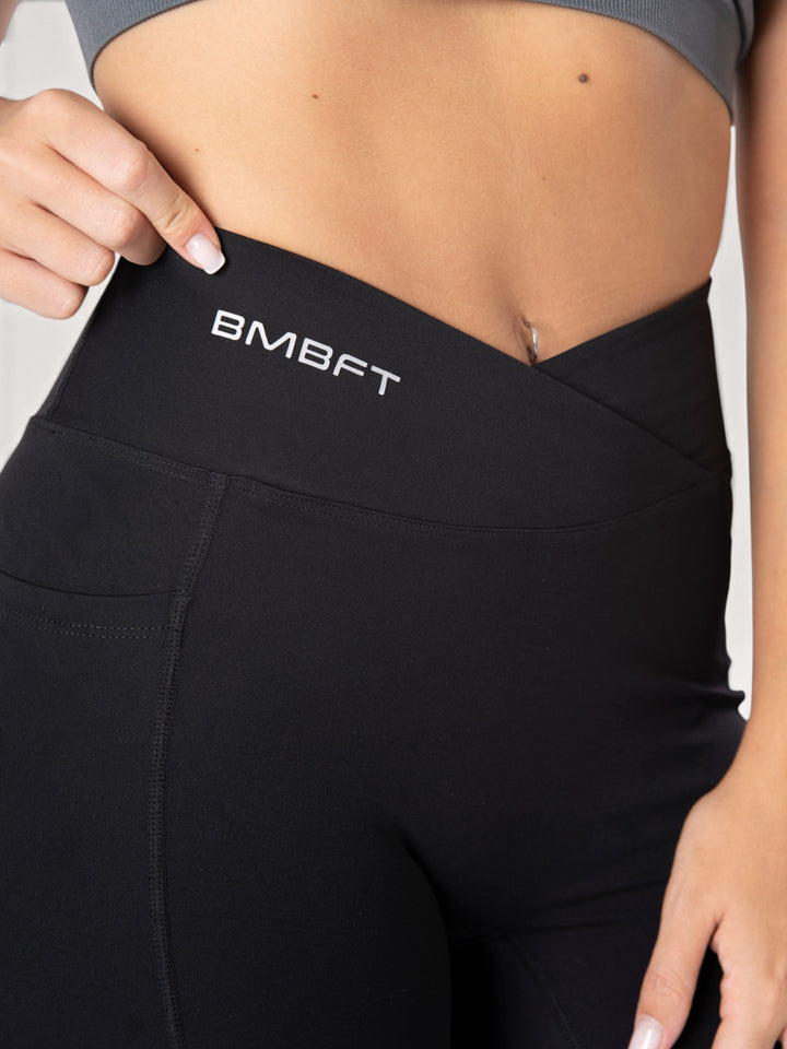 Dettaglio vita alta incrociata dei leggings neri Bombafit con logo stampato e tasca laterale per fitness