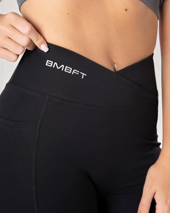 Dettaglio vita alta incrociata dei leggings neri Bombafit con logo stampato e tasca laterale per fitness